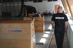 mężczyzna skacze przez skrzynię do ćwiczeń, po prawej stronie obserwuje go instruktor