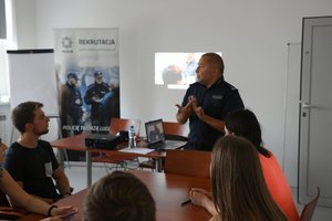 policjant stoi i rozmawia w sali z osobami siedzącymi na krzesłach przy stoliku, w tle baner z napisem policja