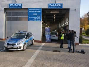 Na zdjęciu budynek stacji diagnostycznej przed którą stoi oznakowany radiowóz policyjny oraz policjantka udzielająca wywiadu ekipie telewizyjnej.