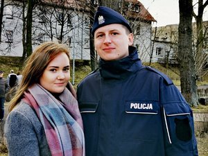 Policjant razem z dziewczyną pozują do zdjęcia, w tle budynek koszarów policyjnych.