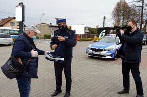 Umundurowany policjant wręcza mężczyźnie odblaskową torbę. Obok nich, niedaleko policyjnego radiowozu stoi operator kamery, który nagrywa materiał wideo dla telewizji.