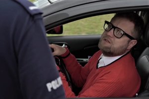 Z lewej strony  widać fragment rękawa policyjnego munduru i znajduje się na nim napis policja. W centralnej części zdjęcia w samochodzie siedzi mężczyzna. Jest to członek kabaretu, a zdjęcie stanowi fragment skeczu z jego udziałem.