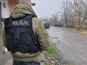 Policjant ubrany w kamizelkę z napisem policja stoi na pierwszym planie, a za nim znajdują się samochody biorące udział w pościgu.
