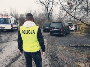 Policjant ubrany w kamizelkę z napisem policja stoi na pierwszym planie, a za nim znajdują się samochody biorące udział w pościgu.