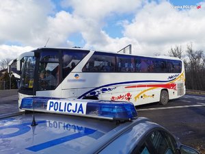 Na zdjęciu widać w oddali zaparkowany autokar koloru białego z kolorowaymi naklejkami. Na pierwszym planie widać radiowóz policyjny. Zdjęcie zrobione w porze dziennej.