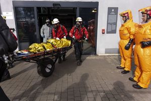 Ratownicy wynoszą poszkodowanego na noszach. Obok stoją mężczyźni w specjalnych, pomarańczowych kombinezonach.