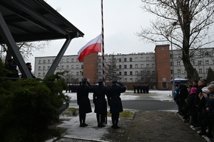 Na zdjęciu widać trzech policjantów stojących tyłem podnoszących Flagę Państwową. Na wprost policjantów przodem do Flagi Państwowej stoją nowo przyjęci policjanci.