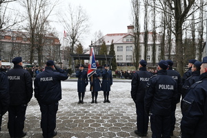 Na zdjęciach widać stojących tyłem policjantów nowo przyjętych w czasie uroczystości ślubowania. Na wprost policjantów przodem stoi trzech policjantów wraz z godłem Państwowym. W tle widać rodziny policjantów nowo przyjętych.