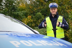 Policjant w odblaskowej kamizelce uśmiecha się, a na pierwszym planie widać fragment radiowozu z napisem Policja.