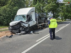 zdjęcie kolorowe przedstawiające umundurowanego policjanta podczas oględzin, a za nim uszkodzony pojazd w wyniku wypadku