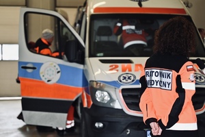 na zdjęciu widoczny jest ambulans medyczny oznakowany z otwartymi drzwiami na wprost niego stoi kobieta w stroju ratownik medycznego. Na plecach kurtki widoczny  jest napis ratownik medyczny