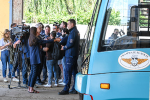 Na zdjęciu widać grupę osób oraz operatora kamery wraz z dziennikarzem, która trzyma mikrofon blisko umundurowanego funkcjonariusza Policji.