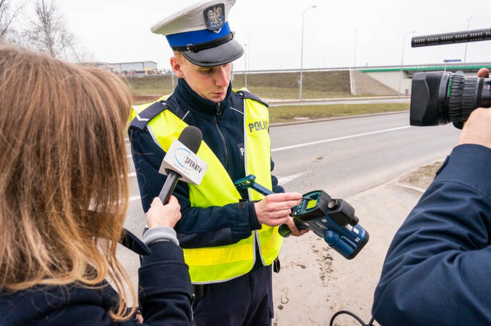 Zdjęcie - po lewej kobieta stojąca bokiem i trzymająca mikrofon w ręku, policjant prezentuje radarowy miernik prędkości, po prawej widoczny obiektyw kamery