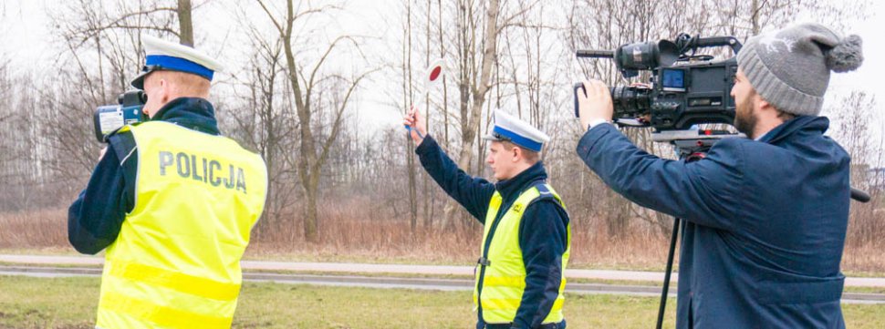 Zdjęcie - dwóch policjantów i operator kamery z kamerą TV na statywie. Jeden policjant dokonuje pomiaru prędkości, a drugi wydał sygnał do zatrzymania pojazdu