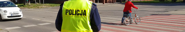 zdjęcie - przejście dla pieszych, biały samochód zatrzymał się aby przepuścić pieszą z rowerem. Na pierwszym planie plecy policjanta w kamizelce odblaskowej.