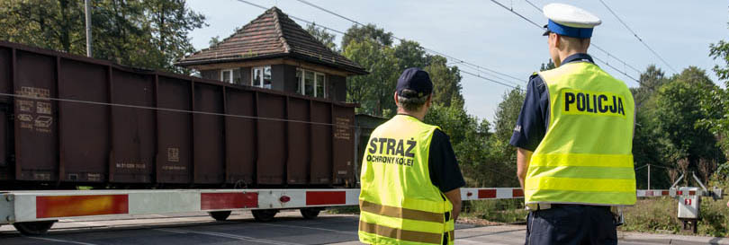 Zdjęcie przedstawia policjanta drogówki i funkcjonariusza SOK przy przejeździe kolejowym w tle wagony 