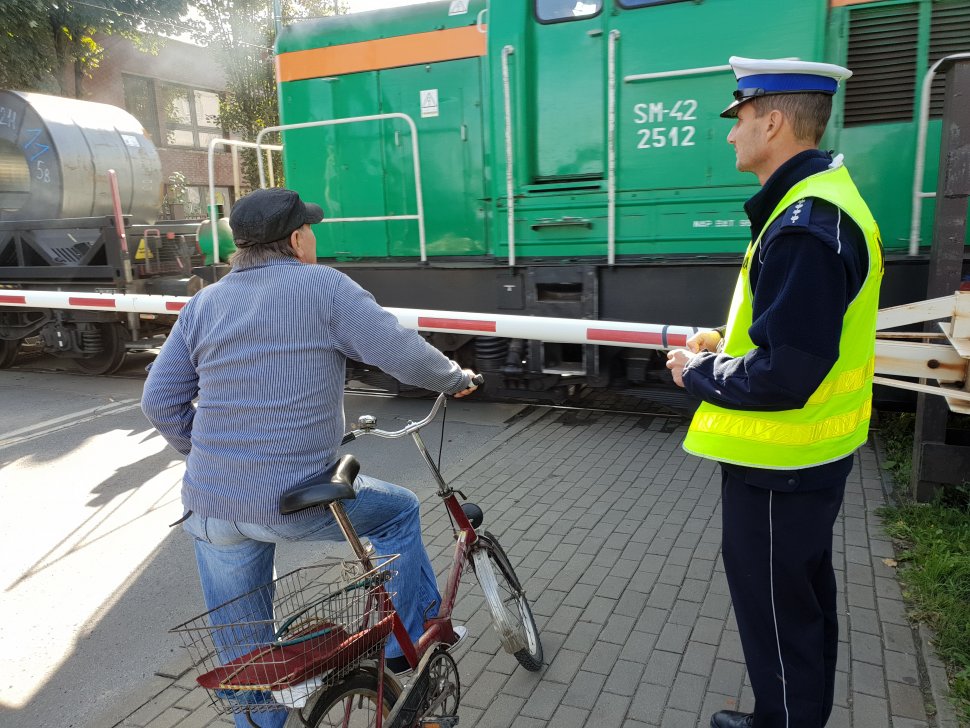 Zdjęcie przedstawia policjanta drogówki i rowerzystę przy przejeździe kolejowym w tle lokomotywa i wagony