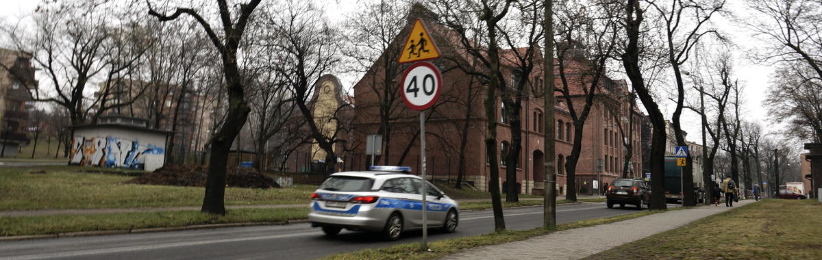 ulica w centrum miasta, jadące samochody i radiowóz policji, znaki ostrzegające o przejściu dla pieszych i dzieciach idących do szkoły