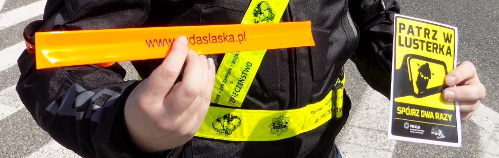 motocyklista trzyma odblaskową opaskę i naklejkę z grafiką i napisem patrz w lusterka spójrz dwa razy
