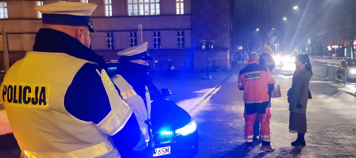 policjanci przy radiowozie, obok ratownik wolontariusz rozdaje przechodniom odblaski