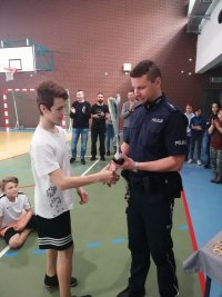 Zdjęcie - hala sportowa, w tle stojące osoby, na pierwszym planie policjant w mundurze wręcza puchar uczestnikowi turnieju.