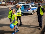 zdjęcie - stojący radiowóz policyjny w tle, na pierwszym planie dwóch chłopców i nauczycielka w żółtych kamizelkach odblaskowych, z prawej strony stoi policjant drogówki