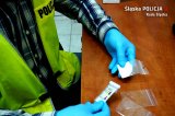 zdjęcie - policjant siedzący przy biurku, w rękach trzyma woreczek z białym proszkiem i tester do badania narkotyków