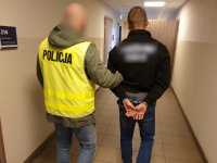 Zdjęcie - policjant w kamizelce odblaskowej z napisem POLICJA i zatrzymany w kajdankach