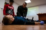 Zdjęcie - policjant w umundurowaniu leży na podłodze, przy nim klęczy strażak i mężczyzna (policjant) po cywilu. Strażak uciska miejsce symulowanej rany.