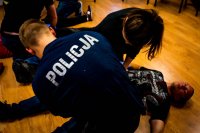 zdjęcie - na podłodze leży mężczyzna, nad nim kobieta i policjant w umundurowaniu - kobieta i policjant uciskają symulowaną ranę