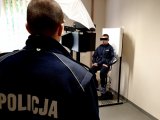 zdjęcie - pomieszczenie techników kryminalistyki - na pierwszym planie tyłem stoi policjant, w tle podejrzany siedzi na stanowisku do robienia zdjęć sygnalitycznych