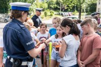 zdjęcie przedstawia - policjantkę rozdającą dzieciom odblaskowe opaski