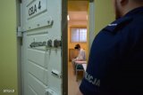 zdjęcie poglądowe - zatrzymany siedzący na pryczy w celi, na zewnątrz policjant