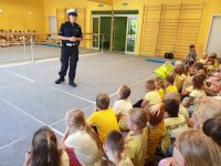 sala gimnastyczna - policjantka stojąca przed siedzącymi pod ścianą dziećmi opowiada o  bezpiecznej drodze do szkołu