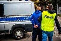 zdjęcie kolorowe - policjant prowadzi zatrzymanego, w tle policyjny radiowóz