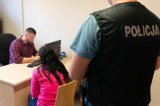 zdjęcie przedstawia pokój przesłuchań, siedzący policjant przesłuchuje zatrzymaną kobietę za którą stoi kryminalny w kamizelce z napisem policja