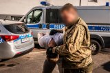 policjant kryminalny prowadzi zatrzymanego, w tle radiowozy oznakowane