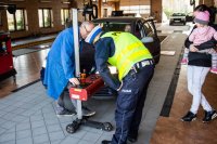 zdjęcie kolorowe - diagnosta i policjant sprawdzają ustawienie świateł w samochodzie, obok kobieta z dzieckiem na ręku
