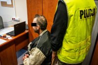 zdjęcie kolorowe, pomieszczenie biurowe, policjant stoi za krzesłem na którym siedzi zatrzymany