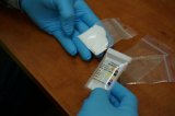 ręce w niebieskich rękawiczkach trzymają tester narkotykowy i woreczek z białym proszkiem