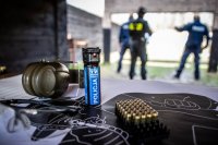 zdjęcie przedstawia zajęcia strzeleckie policjantów - tarcza, miotacz gazu, ochronniki słuchu leżą na stoliku w tle policjanci na stanowisku strzeleckim