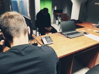 Zdjęcie przedstawia policjanta w cywilu przy biurku, w tle siedzący jeden ze sprawców