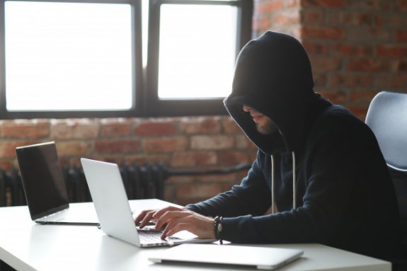 zdjęcie - mężczyzna w kapturze przy stole przed nim dwa laptopy