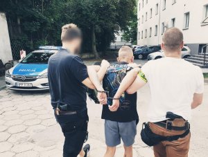 dwaj policjanci po cywilu prowadzą zatrzymanego przez parking w tle samochody i radiowóz
