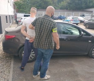 Widok zatrzymanego mężczyzny prowadzonego w kajdankach przez policjanta wydziału kryminalnego.