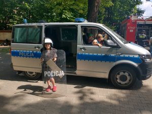 Wizerunek dziecka na tle radiowozu przebranego w policyjny kombinezon przeciwuderzeniowy.
