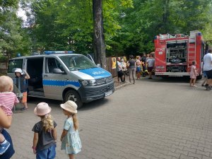 Wizerunek dzieci na tle radiowozu przebranego w policyjny kombinezon przeciwuderzeniowy.