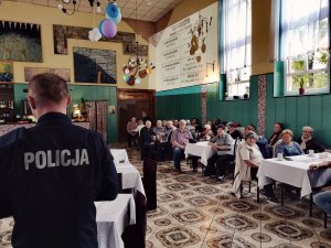 sala z stołami i krzesłami na których siedzą seniorzy, stojący policjant