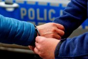 ręka w niebieskiej kurtce, policjant zakłada kajdanki,tło - policyjny radiowóz z napisem policja na drzwiach - zdjęcie poglądowe