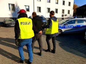 policjanci prowadzą zatrzymanego w tle radiowóz i budynek